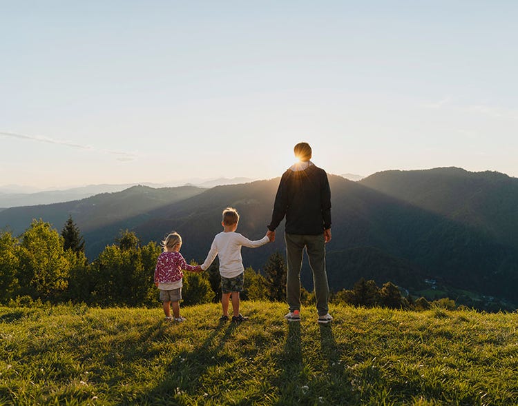 Three people holding hands on a mountain overlook at sunset, facing layered hills and forests under a softly lit sky.